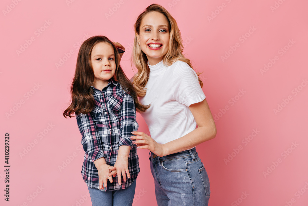 Positive woman in white T-shirt and jeans brushing wavy dark hair of daughter and posing on pink background Positive woman in white T-shirt and jeans brushing wavy dark hair of daughter and posing on pink background
