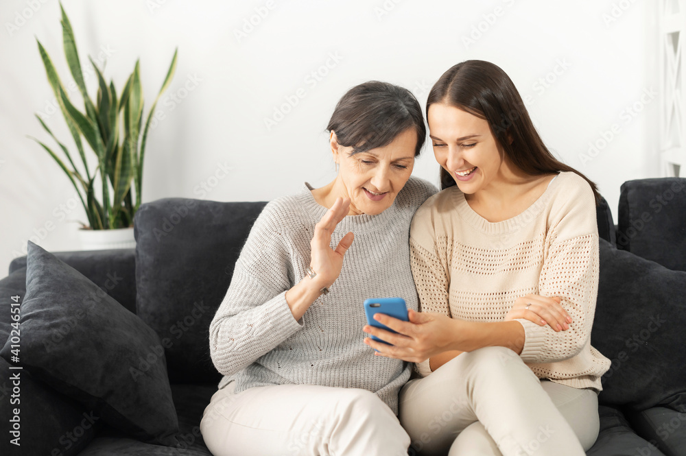 An adult daughter and senior mother using the smartphone for video conference, video call, two multigenerational women sitting on the couch and look at phone webcam and waving hello An adult daughter and senior mother using the smartphone for video conference, video call, two multigenerational women sitting on the couch and look at phone webcam and waving hello