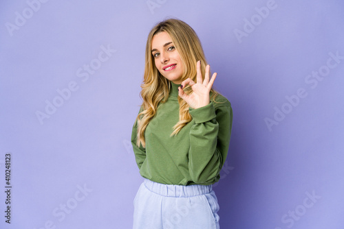 Young caucasian blonde woman winks an eye and holds an okay gesture with hand.