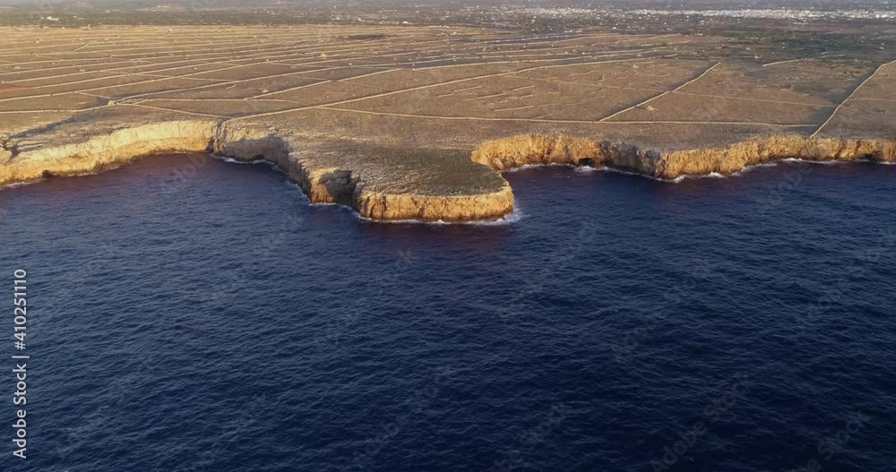 Menorca's coastline with the island in the background, under the sunset. We can see agricultural fields close to the Mediterranean Sea - aerial view with a drone 4K