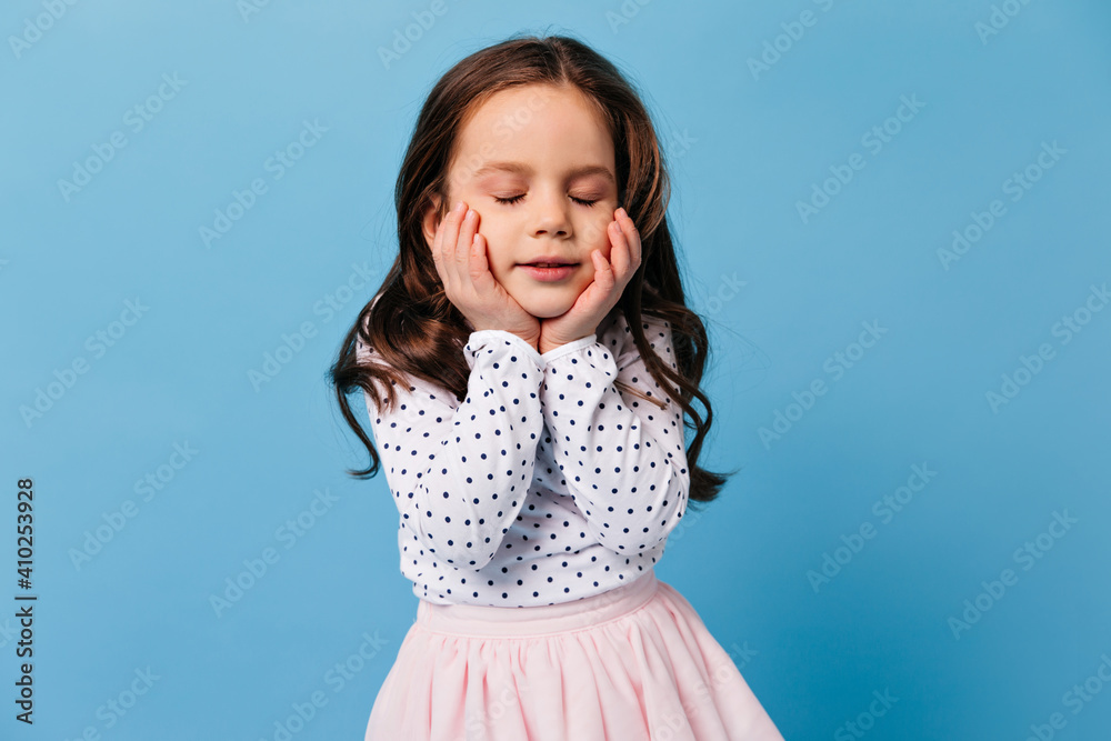 Close-up portrait of dark-haired girl against blue background. Child in polka dot blouse closed her eyes Close-up portrait of dark-haired girl against blue background. Child in polka dot blouse closed her eyes