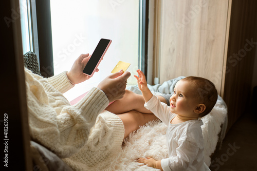 Cute baby girl reaching to credit card being held by mother sitting by window at home photo