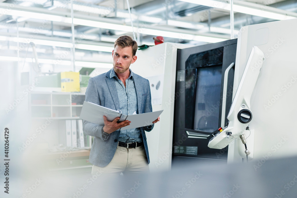 Businessman looking away while holding file document standing by machinery equipment in factory Businessman looking away while holding file document standing by machinery equipment in factory