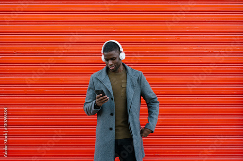 Smiling man wearing headphones using mobile phone while standing against red wall