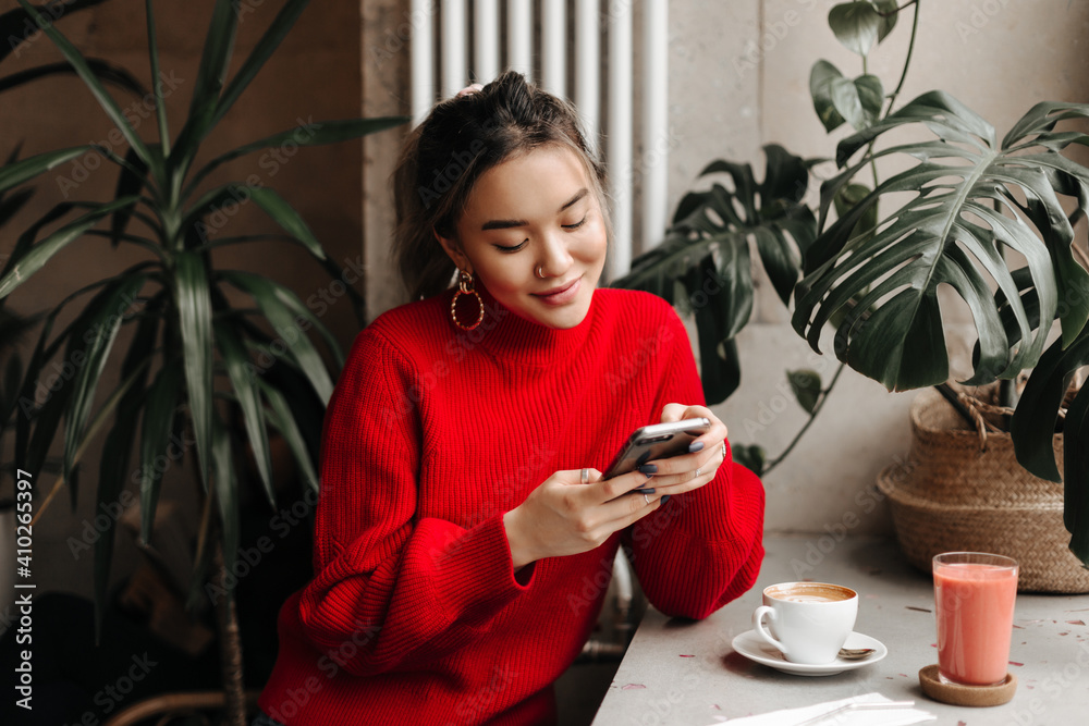 Charming young girl in massive earrings and bright sweater chats in phone while sitting in cafe over cup of coffee - obrazy, fototapety, plakaty 