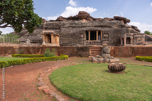 Aihole, Karnataka, India - November 7, 2013: Ravanaphadi Cave Shiva Temple. Black rock with brown stone building additions under blue cloudscape. Green lawn and park in front. photo