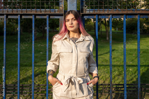 Stylish young woman with hands in pockets against fence on sunny day