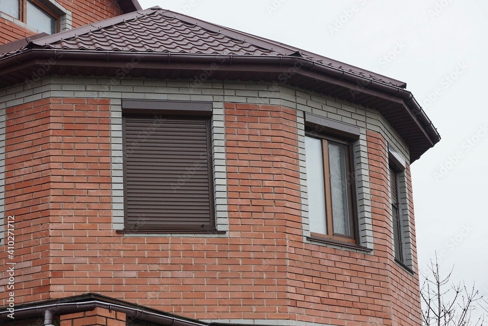 brown brick attic of a private house with windows closed with roller shutters under a tiled roof against a gray sky