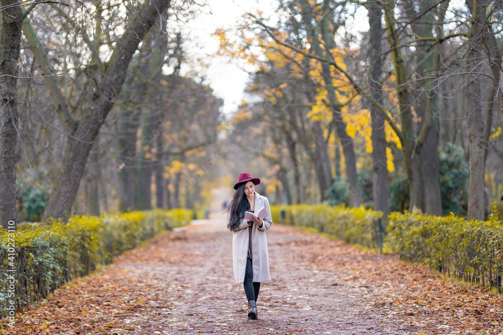 Mid adult woman reading book while walking on land amidst trees in park Mid adult woman reading book while walking on land amidst trees in park
