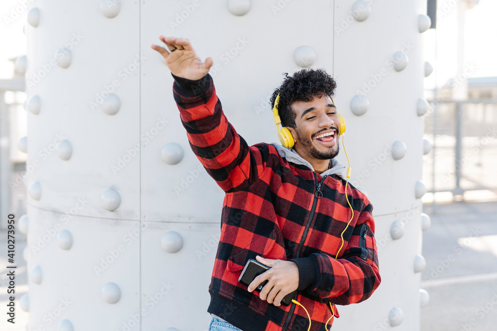 Happy man with mobile phone listening music through headphones while dancing against wall