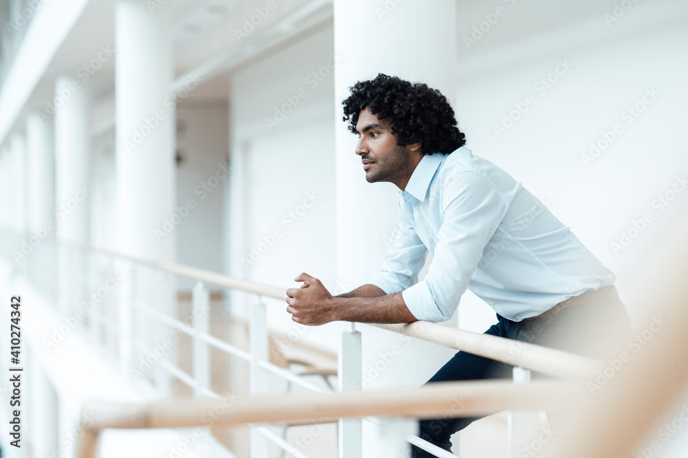Thoughtful young male entrepreneur with black curly hair leaning on railing at office corridor Thoughtful young male entrepreneur with black curly hair leaning on railing at office corridor
