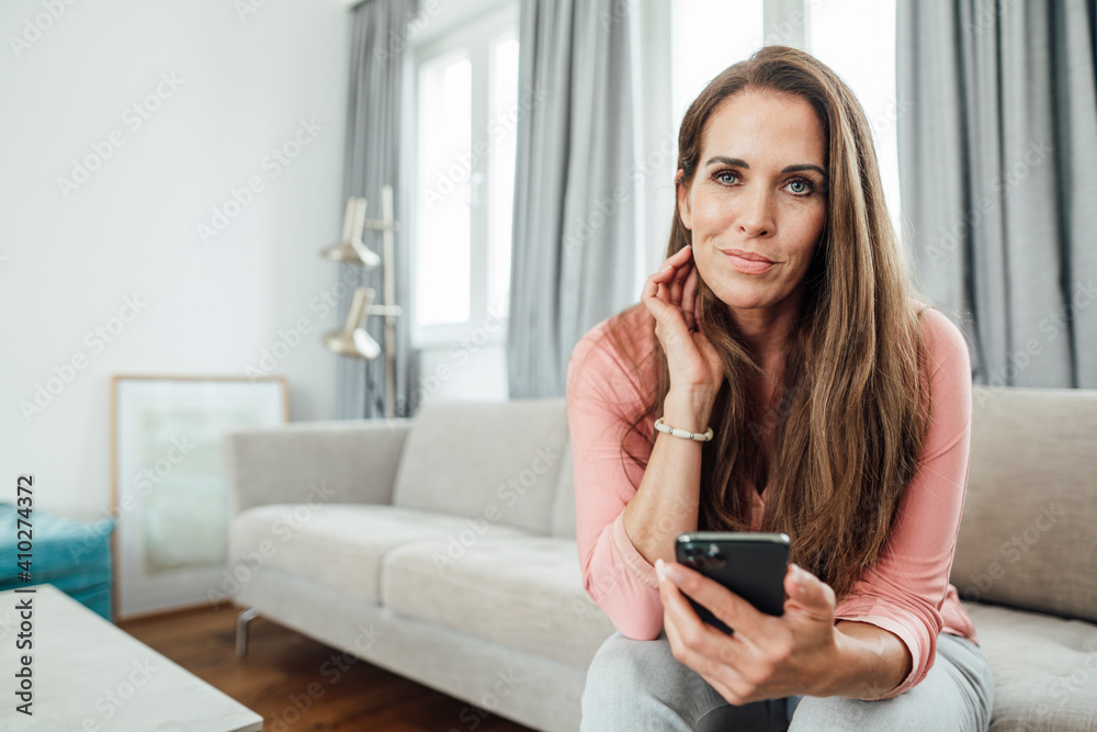 Mature woman with hand in hair holding mobile phone in living room at home Mature woman with hand in hair holding mobile phone in living room at home