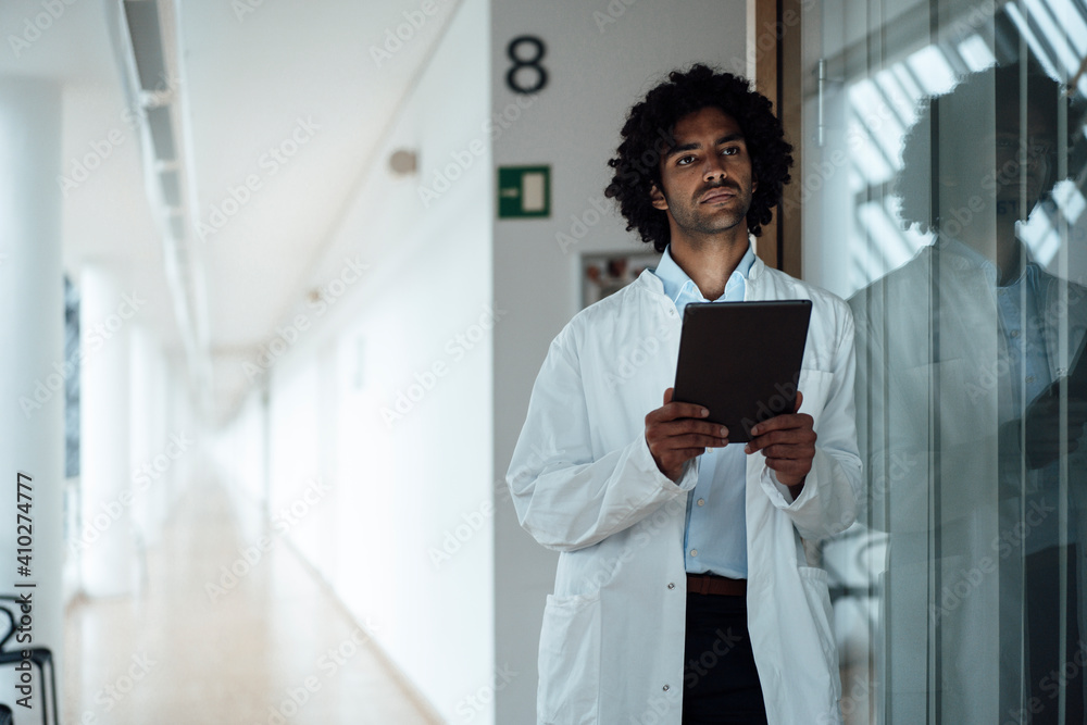 Thoughtful young male doctor holding digital tablet while leaning on glass wall at hospital corridor Thoughtful young male doctor holding digital tablet while leaning on glass wall at hospital corridor