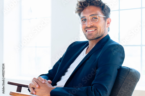 Confident businessman wearing eyeglasses sitting with hands clasped on armchair at home photo