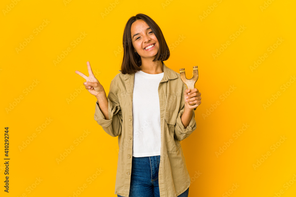 Young hispanic woman holding a slingshot joyful and carefree showing a peace symbol with fingers. Young hispanic woman holding a slingshot joyful and carefree showing a peace symbol with fingers.