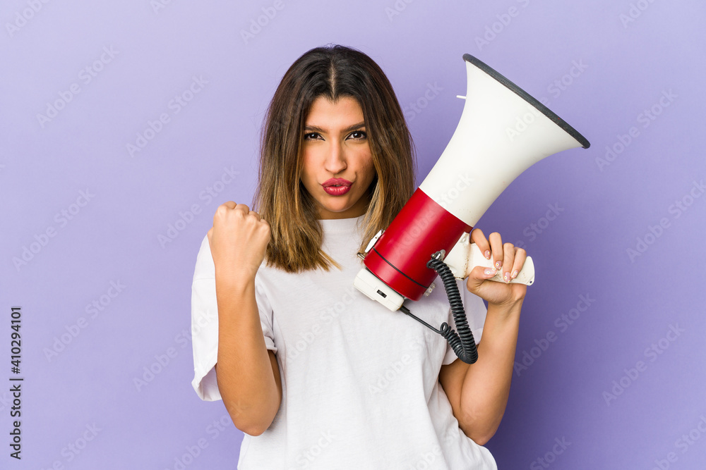 Young indian woman holding a megaphone isolated showing fist to camera, aggressive facial expression. Young indian woman holding a megaphone isolated showing fist to camera, aggressive facial expression.