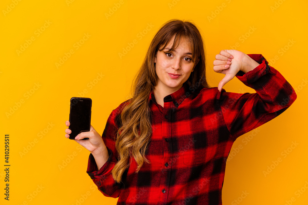 Young caucasian woman holding phone isolated on yellow background showing a dislike gesture, thumbs down. Disagreement concept. Young caucasian woman holding phone isolated on yellow background showing a dislike gesture, thumbs down. Disagreement concept.