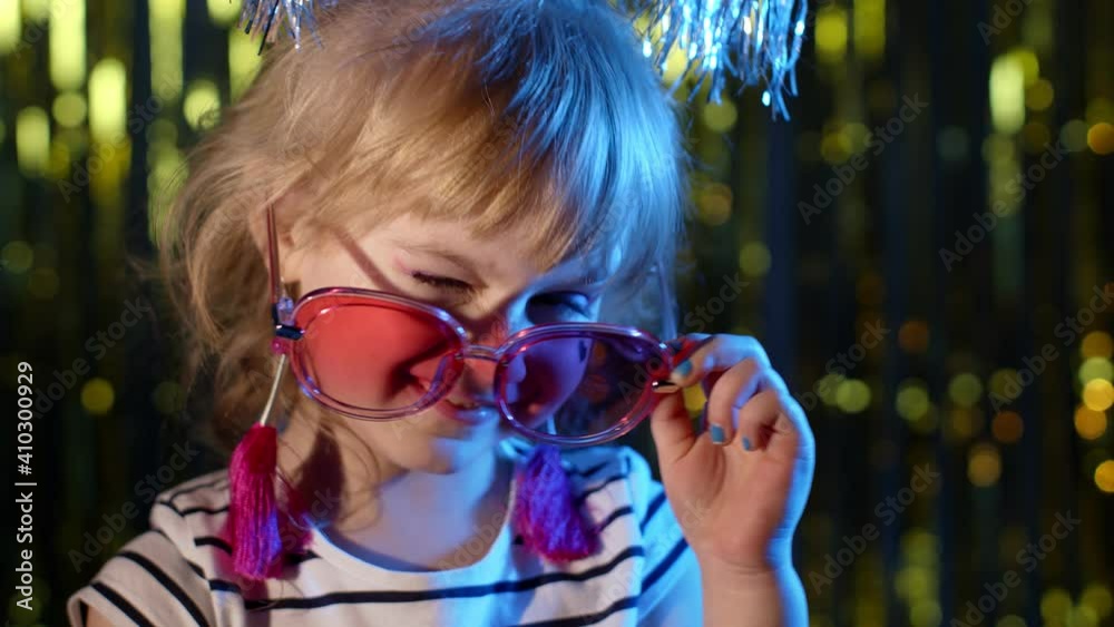 Stylish trendy child kid in red sunglasses at disco party cyberpunk club looking at camera and smiling. Close-up portrait of young blond girl in bright colorful blue neon light on shiny background Stylish trendy child kid in red sunglasses at disco party cyberpunk club looking at camera and smiling. Close-up portrait of young blond girl in bright colorful blue neon light on shiny background