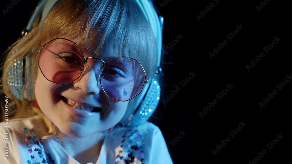 Stylish trendy child kid in sunglasses at disco party cyberpunk club looking at camera and smiling. Close-up portrait of young playful blond girl in bright colorful neon rays light on black background Stylish trendy child kid in sunglasses at disco party cyberpunk club looking at camera and smiling. Close-up portrait of young playful blond girl in bright colorful neon rays light on black background
