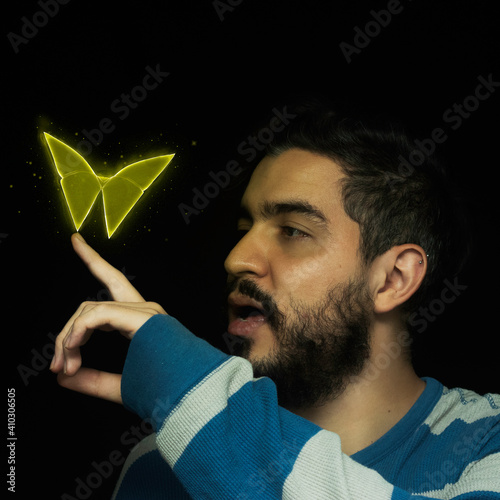 Fondo negro con un hombre joven de buso a rayas azul con blanco, barba y cabello negro mira feliz en la punta de su dedo una mariposa amarilla hecha en origami mágico volando con destellos de luz photo