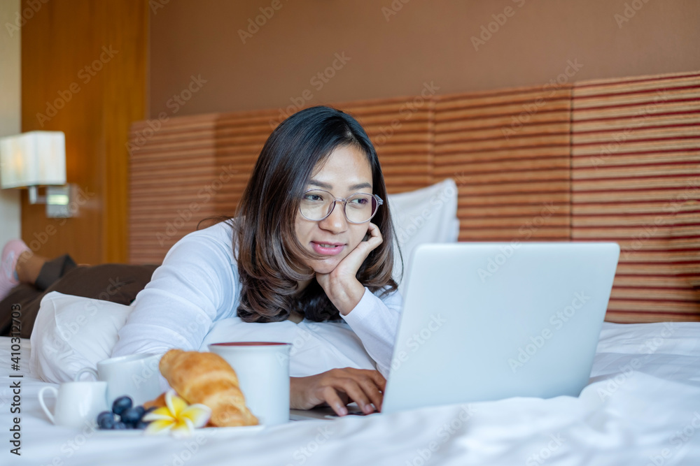 Young Asian women smiling happily at freelance work, working on a notebook while relaxing in bed with snacks and fruit in a hotel room. Vacation and relaxation Young Asian women smiling happily at freelance work, working on a notebook while relaxing in bed with snacks and fruit in a hotel room. Vacation and relaxation