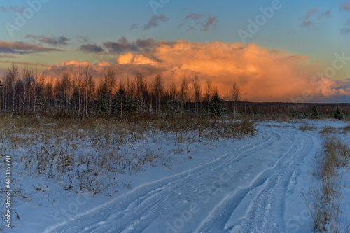 Winter landscape with ruts leading to the horizon, tall trees in an open area and a beautiful sunset sky with a large cloud in the form of a heap, painted in orange shades. Frosty winter sunny day 