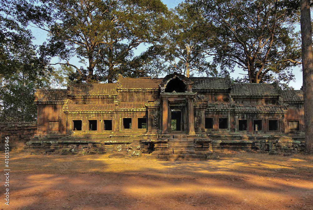 Obraz premium Ancient stone palace in Angkor. Empty window and doorways, columns at the entrance are visible. The building is dilapidated and weathered. Tropical trees against the blue sky. Cambodia