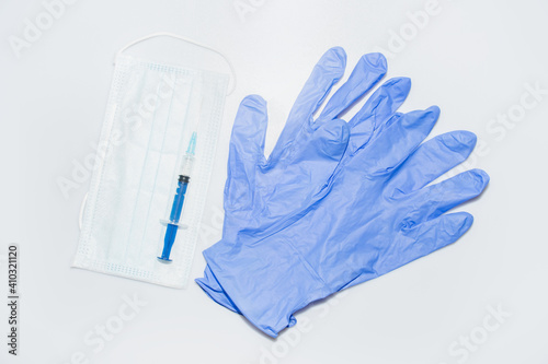 A pair of blue latex gloves, a protective face mask, and a syringe with a medicine or vaccine on a white or gray background: coronavirus flu epidemic, medical background, top view