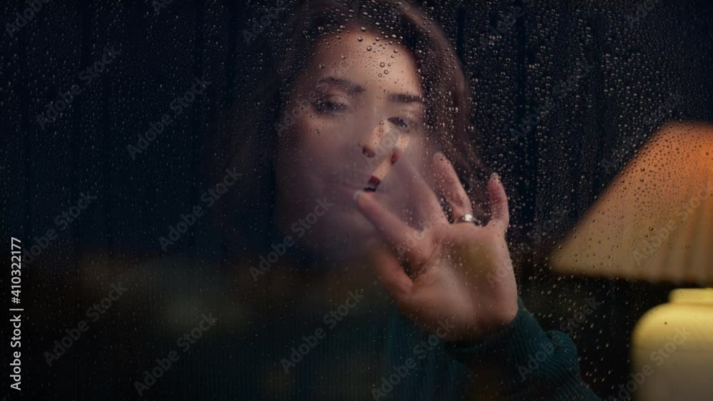 Young woman pears out at rainy night, breathes on glass  and draws a heart on window