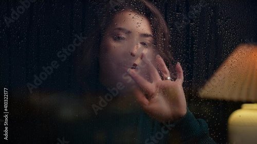 Young woman pears out at rainy night, breathes on glass  and draws a heart on window
