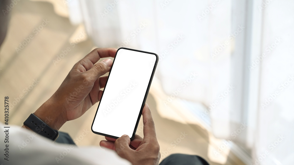 Close up view of young man sitting near window and holding mock up smart phone with white screen. Close up view of young man sitting near window and holding mock up smart phone with white screen.