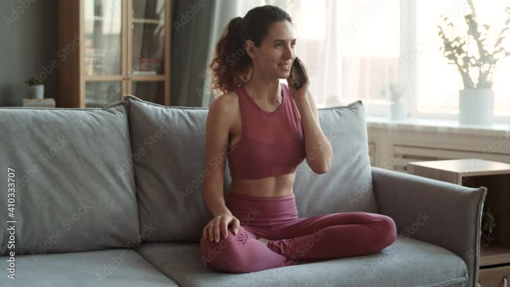 Full shot of young beautiful Caucasian woman wearing yoga pants and crop top, sitting on couch in lotus position, talking on mobile phone and smiling Full shot of young beautiful Caucasian woman wearing yoga pants and crop top, sitting on couch in lotus position, talking on mobile phone and smiling