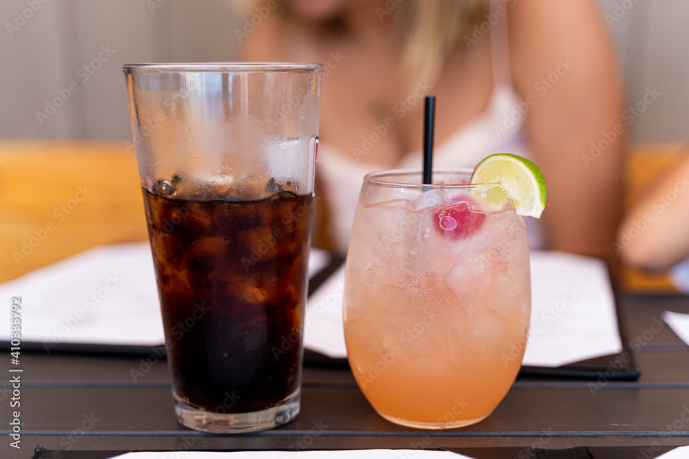 Cup of pink drink, fresh lemonade, with cherry and lemon and straw next to cup of soda full of ice, woman out of focus in background, authentic feel Cup of pink drink, fresh lemonade, with cherry and lemon and straw next to cup of soda full of ice, woman out of focus in background, authentic feel