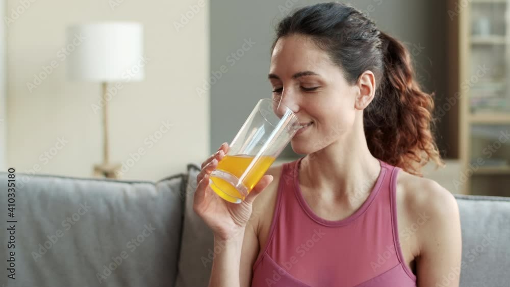 Chest-up of young cheerful sporty Caucasian woman wearing hair up in ponytail, sitting on couch in living room, enjoying drinking orange-colored juice from glass, smiling Chest-up of young cheerful sporty Caucasian woman wearing hair up in ponytail, sitting on couch in living room, enjoying drinking orange-colored juice from glass, smiling