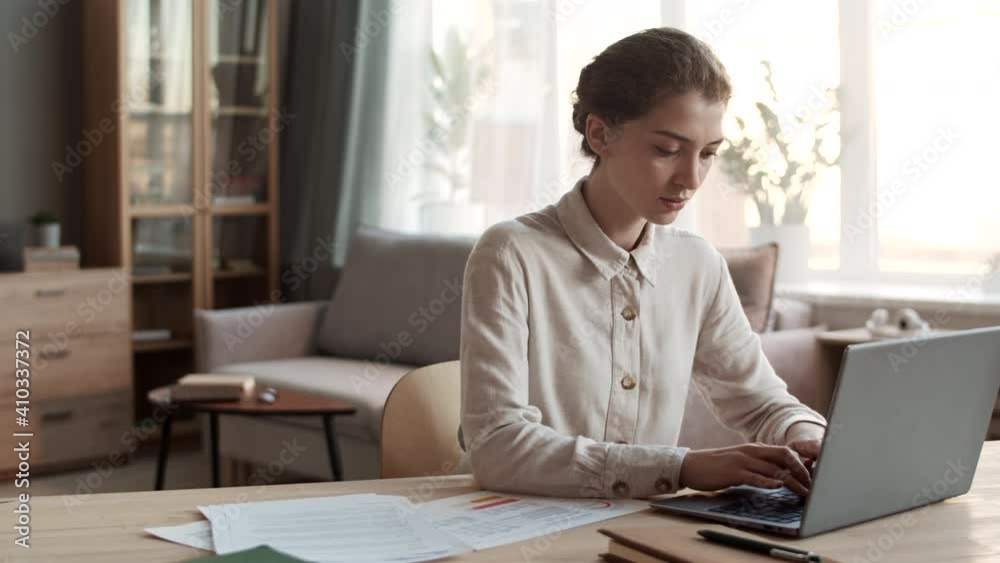 Medium close-up of young professional Caucasian woman wearing formal shirt, sitting by desk in home office, typing on laptop computer, then looking at documents, working from home Medium close-up of young professional Caucasian woman wearing formal shirt, sitting by desk in home office, typing on laptop computer, then looking at documents, working from home