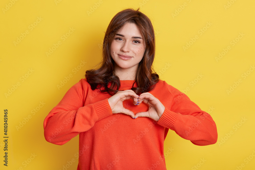 Fototapeta premium Charming young girl showing heart gesture, smiles happily, has romantic feelings, seeks lonely hearts, feels passion, dresses in casual orange sweater, posing isolated over yellow background.