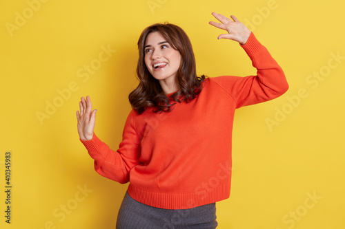 Winsome young European female in orange jumper and grey leggins, looking smiling aside, keeping hands up, standing isolated over yellow background, celebrating success.