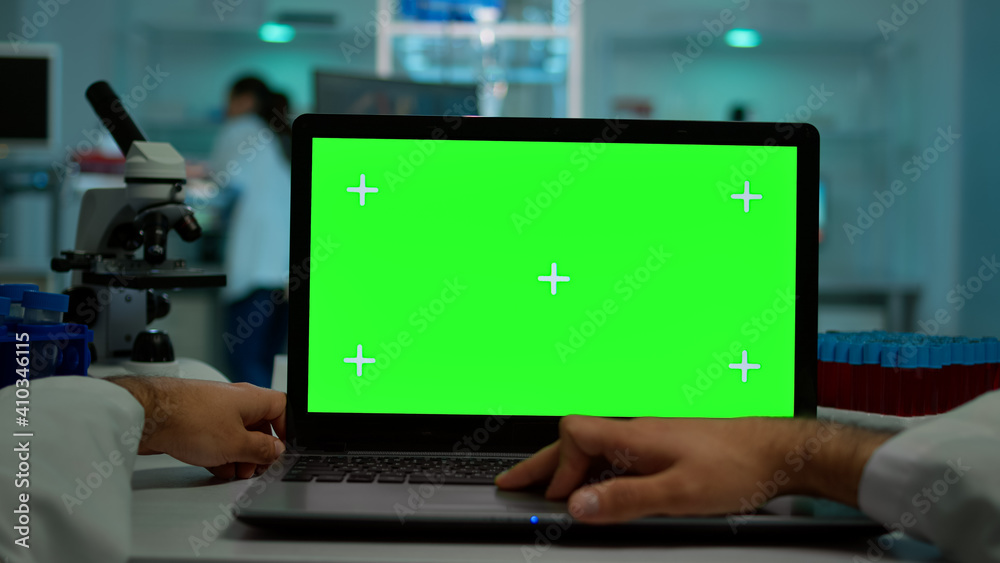 Pov shot of man scientist sitting at desk working on laptop with mock-up green screen, isolated display. In background lab researcher analysing vaccine developent examining samples
