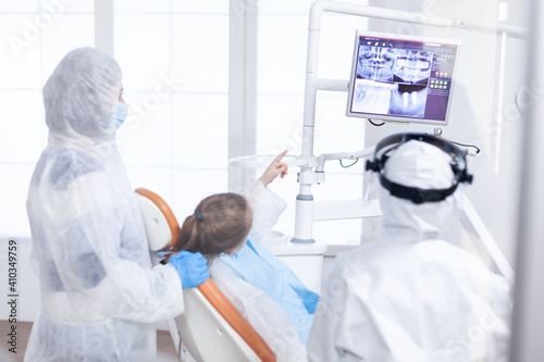 Little girl in ppe suit pointing at digital radiography during consultation. Stomatolog in protectie suit for coroanvirus as safety precaution looking at digital child teeth x-ray during consultation. photo