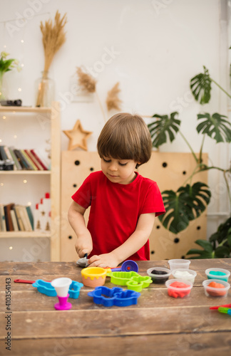 small toddler boy in a red T-shirt plays with colorful plasticine at a wooden table in the room. Development of fine motor skills
