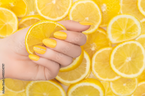 Female hand with yellow nail design. Yellow nail polish manicure. Woman hand with yellow lemons background. Copy space.