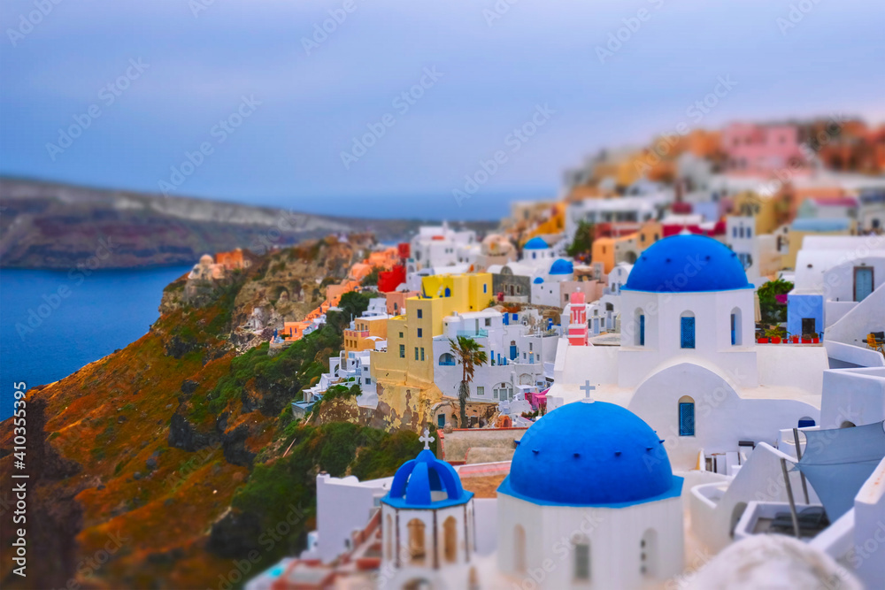 Famous greek iconic selfie spot tourist destination Oia village with traditional white houses and church in Santorini island on sunset in twilight, Greece. Toy camera tilt shift miniature effect