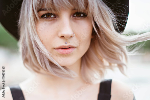 Close up portrait of blonde hipster woman in black hat and dress with tattoos and piercing outdoors