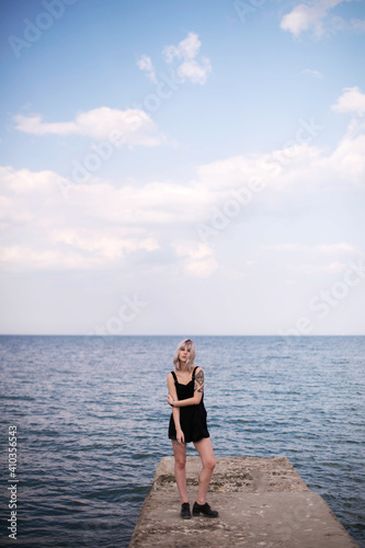 Attractive blonde hipster girl with tattoos wearing black dress standing on the concrete pier on the sea. Outdoors © WellStock