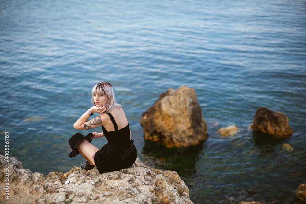 Beautiful young blonde hipster girl with tattoos wearing black dress, holding black hat sitting on the stony beach near the sea. Outdoors
