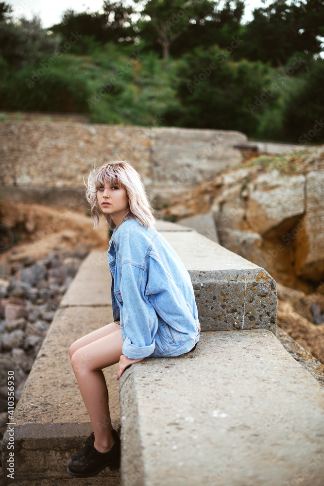Blonde hipster girl with blonde hair in jeans jacket sitting outdoors on the rocks. Blonde hipster girl with blonde hair in jeans jacket sitting outdoors on the rocks.