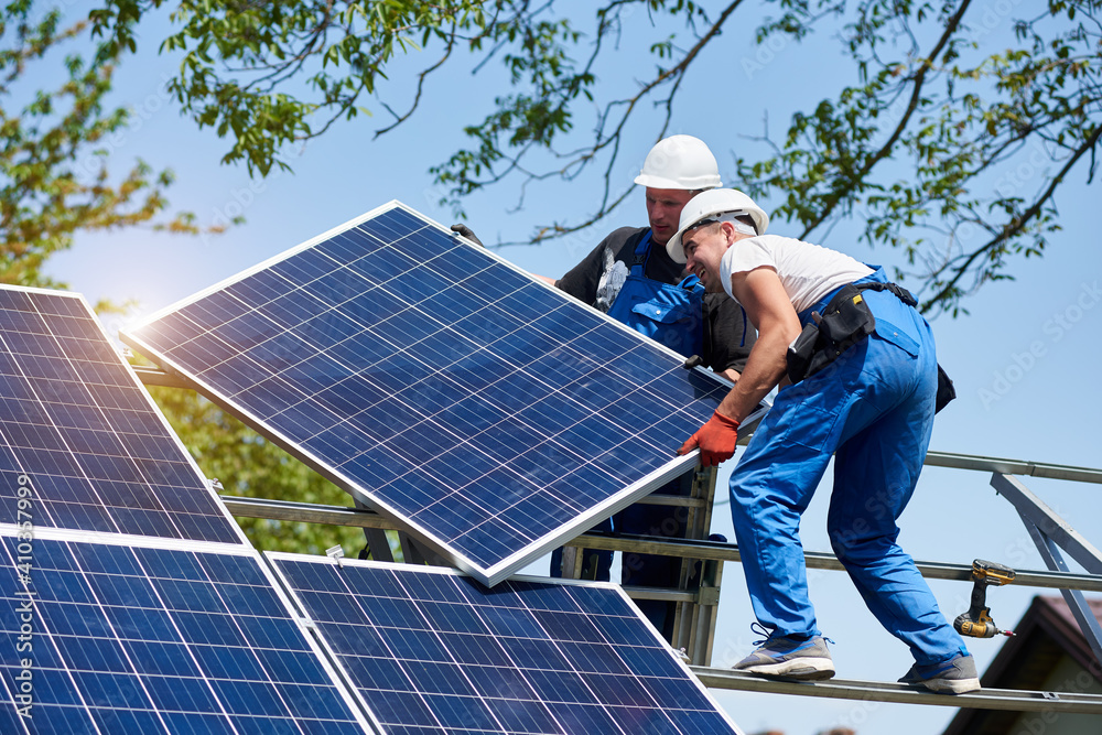 Two young technicians mounting heavy solar photo voltaic panel on tall steel platform on green tree background. Exterior solar panel voltaic system installation, dangerous job concept. Two young technicians mounting heavy solar photo voltaic panel on tall steel platform on green tree background. Exterior solar panel voltaic system installation, dangerous job concept.