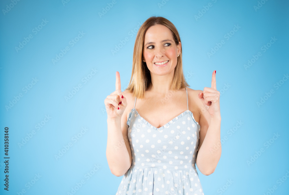 Portrait of a happy young woman wearing casual dress pointing finger up at copy space isolated over blue background Portrait of a happy young woman wearing casual dress pointing finger up at copy space isolated over blue background