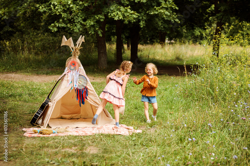 Funny blonde girls playing next to wigwam in the forest. Children games, leisure time. © Natalia