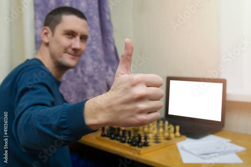 Mock-up online chess courses for adults. A man shows the class by raising his thumb up against the background of a chessboard and a laptop with a place for a layout. Close-up. Copyspace